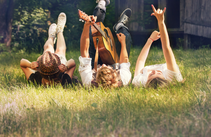 3 jongvolwassenen liggen in het gras, een heeft een gitaar, de zon schijnt.
